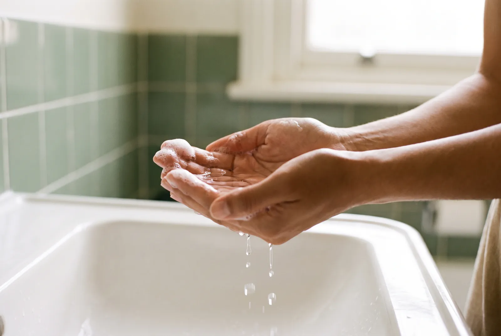 Manos juntas recogiendo agua sobre un lavamanos de cerámica, con gotas que caen y un poco de espuma suave