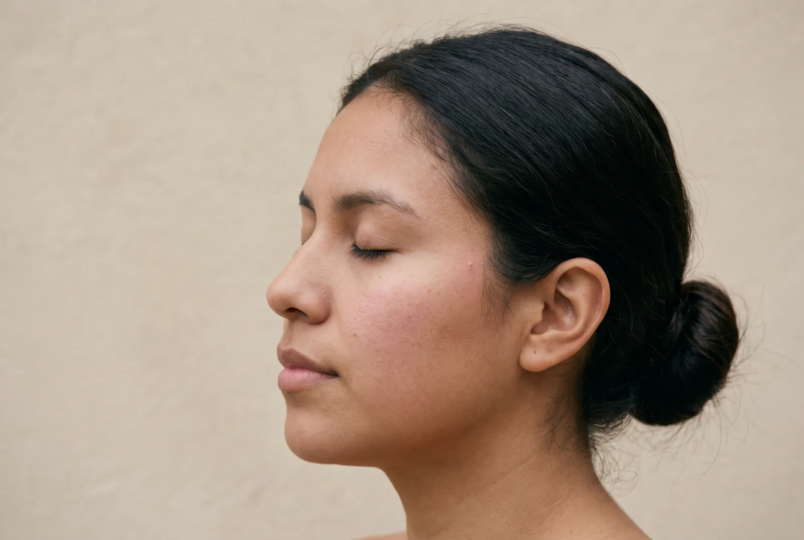 Perfil de una mujer de piel trigueña con los ojos cerrados y un leve rubor natural en la mejilla, bajo una luz suave