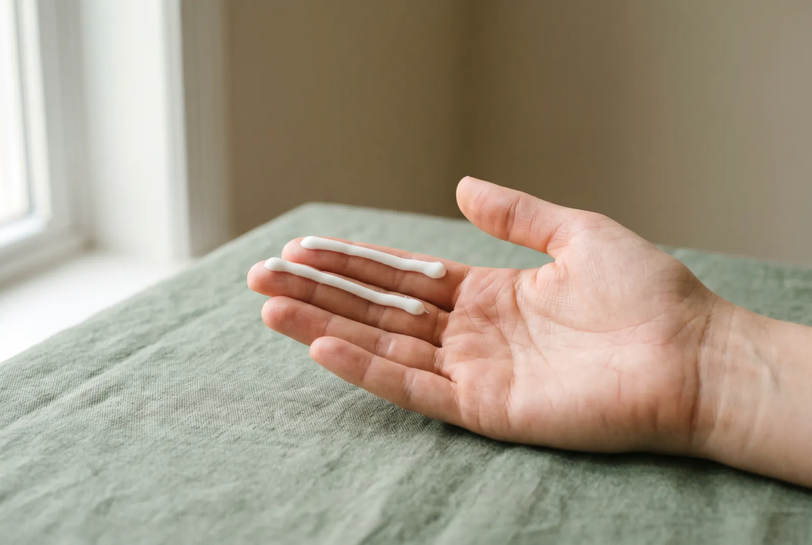 Dos líneas de protector solar blanco a lo largo de los dedos índice y corazón de una mano, con luz de ventana y fondo verde salvia