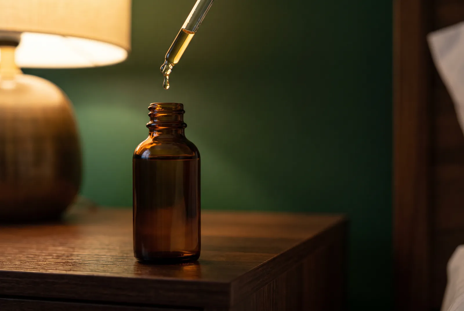 Frasco de vidrio ámbar en una mesa de noche de madera, con un gotero que deja caer una gota de sérum dorado junto a una lámpara encendida